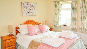 Una cama bien hecha con ropa de cama rosa y blanca, una mesita de noche de madera con una lámpara y una ventana con cortinas estampadas.