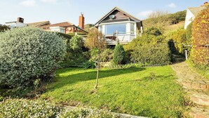 A house with a large glass window, a wooden deck, and a garden with various plants and a lawn. - Bonito is situated in Ventnor with panoramic sea views. (Ventnor)