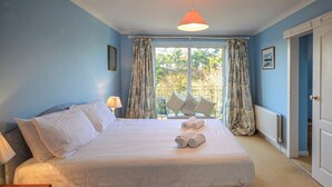 Una cama bien tendida con sábanas y toallas blancas, una lámpara de noche y vistas al exterior a través de una ventana con cortinas.