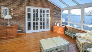 Habitación iluminada por el sol con piso de madera, puerta de cristal que da al exterior, mobiliario de mimbre y vistas amplias a través de grandes ventanales.