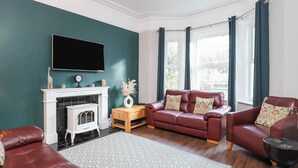 Una sala de estar con chimenea, un sofá, un sillón de cuero rojo, un televisor y una ventana con cortinas.