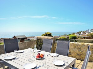 An outdoor dining table with chairs, plates, cutlery, and a bottle of water, set against a backdrop of a coastal view. - 2 bedroom accommodation in Ventnor (Ventnor)
