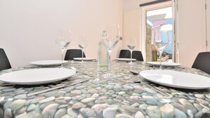 Een eettafel met een glazen blad, omringd door stoelen, borden en wijnglazen, met een raam en een deur op de achtergrond.
