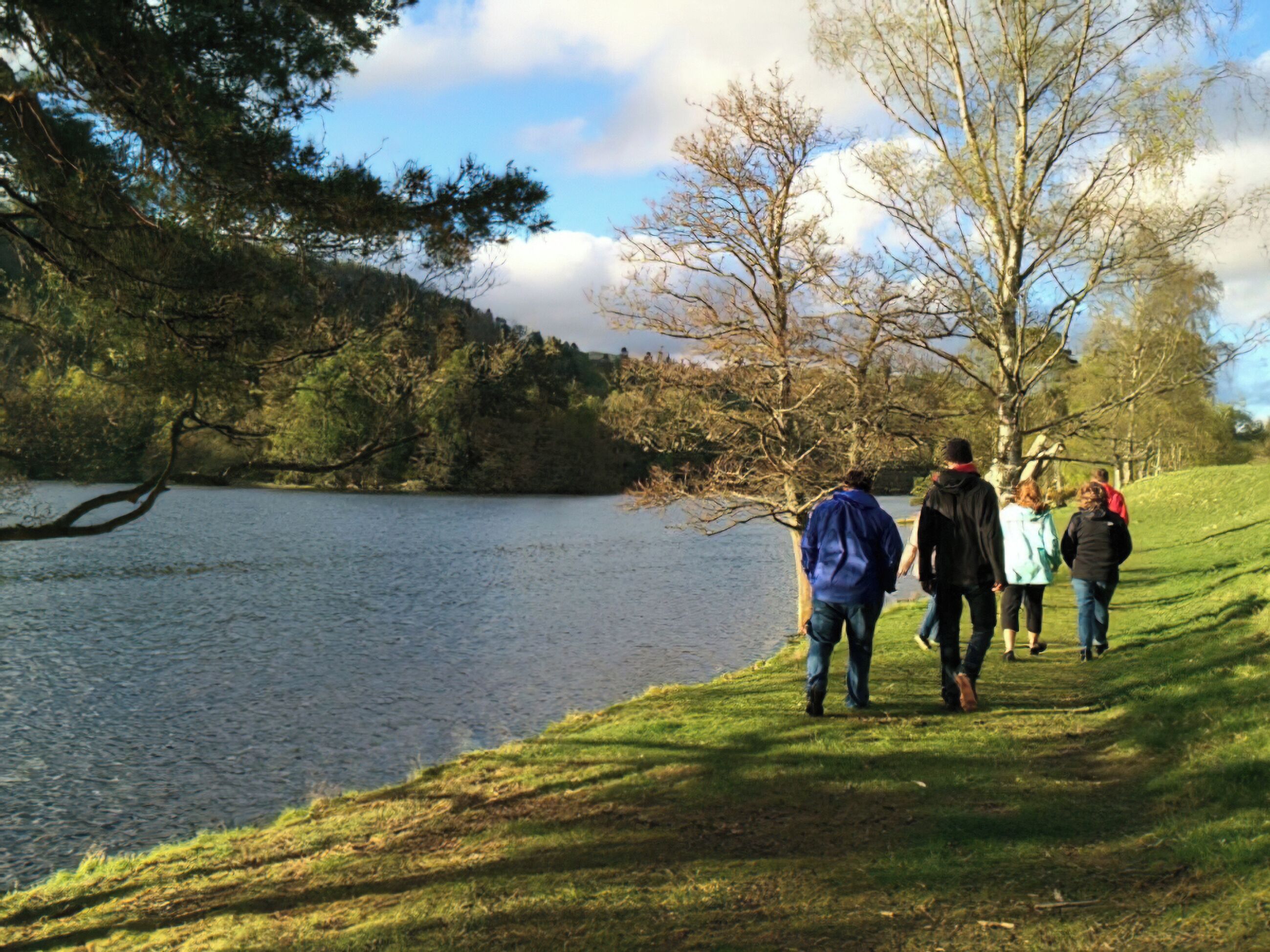 Un groupe de personnes se promène le long d’un sentier bordé d’herbe, au bord d’un lac, entouré d’arbres.