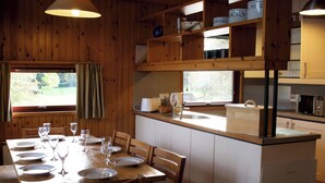 Un comedor de madera con una mesa preparada para cuatro personas, una cocina con armarios blancos y un ventanal con cortinas.