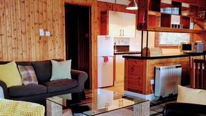 A cozy living room with a sofa, a glass coffee table, and a wicker chair. A kitchen with wooden cabinets and a refrigerator is visible in the background. - Cedar Chalet 20 - UKC5238 (Crieff)