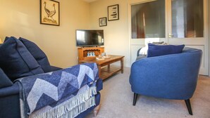 Een woonkamer met een bank, een blauwe fauteuil, een televisie op een houten dressoir en fotolijstjes aan de muur.