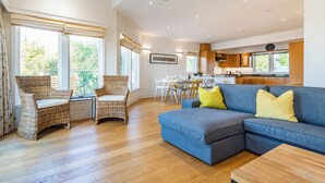 A spacious living area with a blue sofa, wicker chairs, a dining table, and a kitchen in the background.