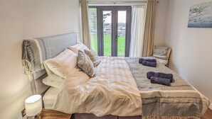 Una cama bien tendida con cabecera gris, una lámpara de noche y vista a un jardín verde a través de una puerta de vidrio.