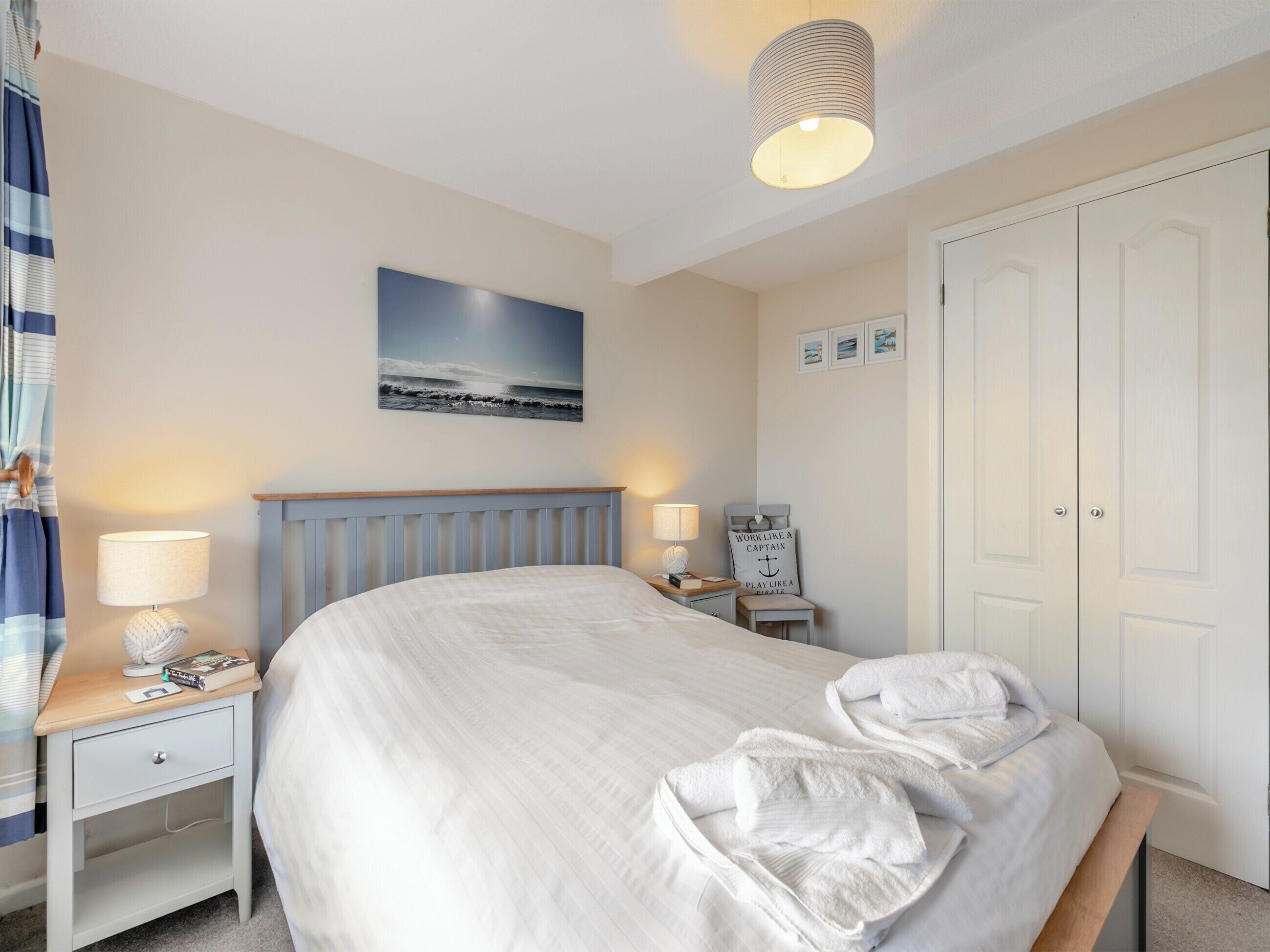 Un lit bien fait, avec du linge de lit blanc, une table de chevet avec une lampe, un tableau accroché au mur et une porte dotée d’un miroir.