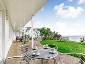 Un patio cubierto con una mesa preparada para dos, con vistas a una zona costera con casas y vegetación.