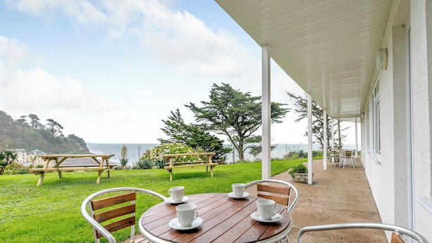 Een overdekt terras met een houten tafel en stoelen, met uitzicht op een gazon en de zee.