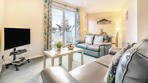 Un salon doté d’un téléviseur à écran plat, d’un canapé, d’une table basse agrémentée d’une plante, et offrant une vue sur la mer grâce à de grandes fenêtres.