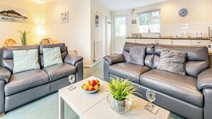 Un salon moderne avec deux canapés en cuir gris, une table basse sur laquelle se trouve un saladier rempli de fruits et une plante, et une cuisine en arrière-plan.