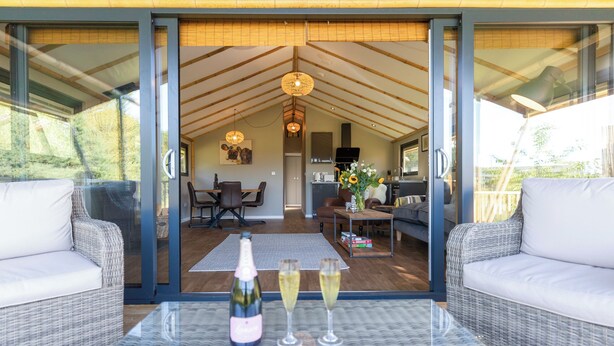 A modern living room with a glass partition, wicker furniture, a dining area with a table and chairs, and a kitchen in the background. - 2 bedroom accommodation in Stanton-On-The-Wolds (Stanton-On-The-Wolds)