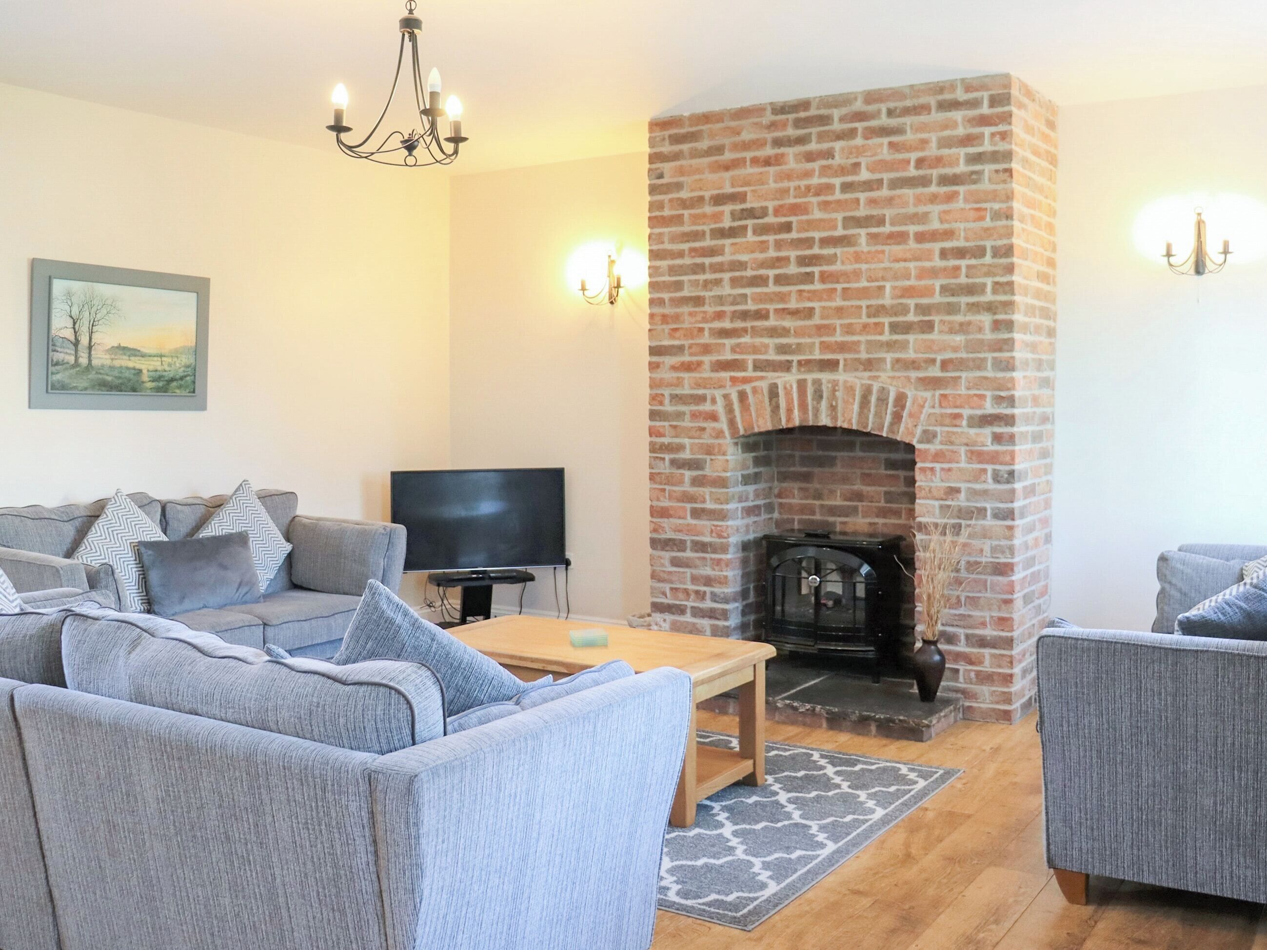 Ein Wohnzimmer mit Kamin aus Backstein, grauen Sofas, einem Fernseher und einer Chandelier.
