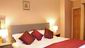 A neatly made bed with a red blanket and pillows, a bedside table with a lamp, and a wooden door in the background. - 4 bedroom accommodation in Freshwater East near Pembroke (Freshwater East near Pembroke)