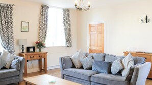 A living room with a grey sofa set, a wooden coffee table, patterned curtains, a framed picture, and a chandelier. - 4 bedroom accommodation in Freshwater East near Pembroke (Freshwater East near Pembroke)