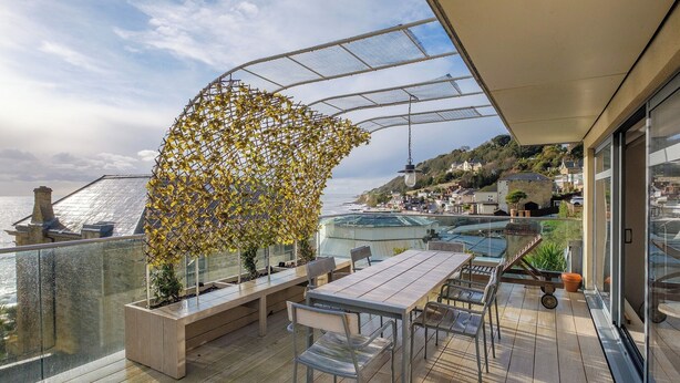 Eine Holzterrasse mit Tisch und Stühlen, einem Glasgeländer und Blick auf das Meer und Gebäude.