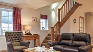Un salon avec un escalier en bois, deux fauteuils, une table basse avec une théière et des tasses, et une fenêtre avec des rideaux.