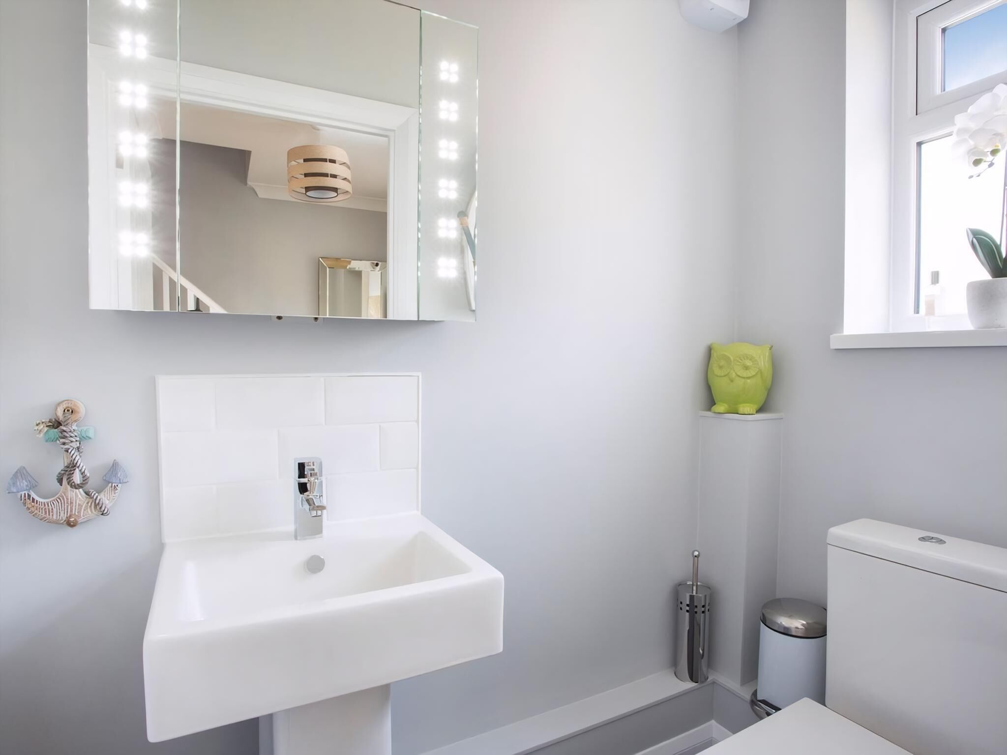 A bathroom with a white sink, a mirror with lights, a toilet, and a window with a plant.