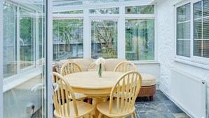 Ein Wintergarten mit einem runden Holztisch und Stühlen, einer Couch und großen Fenstern mit Blick auf grüne Bepflanzung.