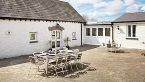 Un bâtiment blanc avec un toit recouvert de tuiles, un patio abrité avec une table et des chaises, et une porte dotée de panneaux de verre.