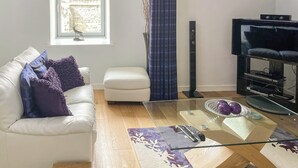 A living room with a white sofa, a glass coffee table, a television on a stand, and a window with a view of buildings. - Abbey Church 14 - UKC5423 (Fort Augustus)