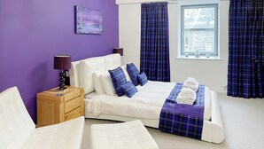 A modern bedroom with a bed, two white chairs, a wooden nightstand, a purple wall, and a window with curtains. - Abbey Church 14 - UKC5423 (Fort Augustus)