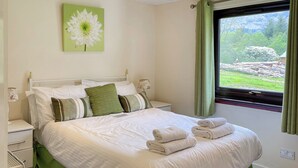 Una cama bien hecha con sábanas blancas y detalles en verde, una mesita de noche con lámpara y una ventana con cortinas.