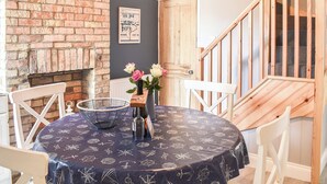 A dining area with a brick fireplace, a round table with a nautical-themed tablecloth, white chairs, and a wooden staircase. - 2 bedroom accommodation in Pakefield (Pakefield)