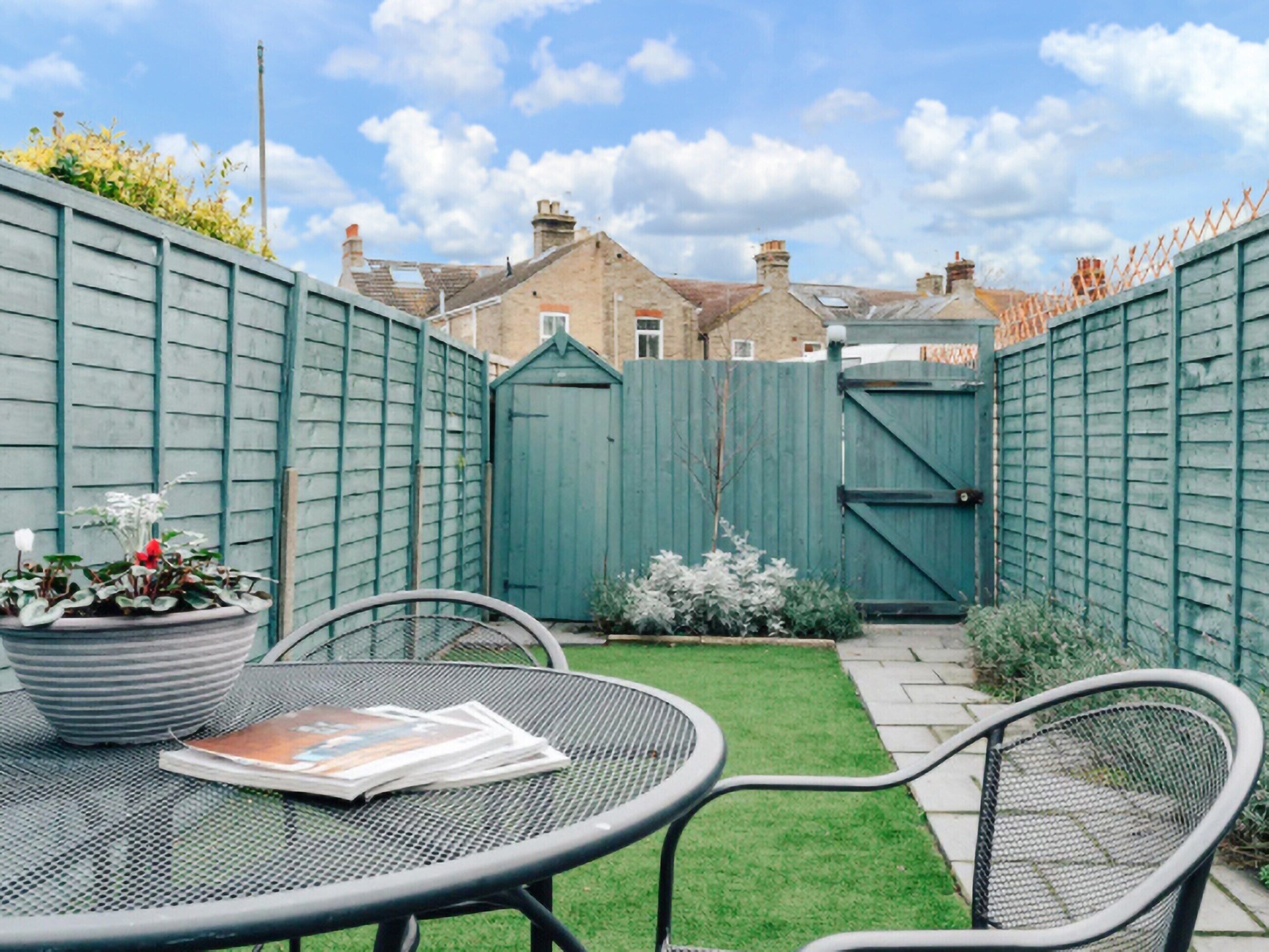 A patio with a metal table and chairs, a small garden area with plants, and a wooden gate in the background.