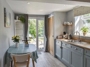 A kitchen with a dining area, featuring a table with a vase of flowers, a coffee maker, and a window with curtains. - 1 bedroom accommodation in Pakefield (Pakefield)