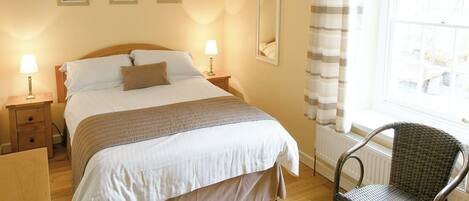 A neatly made bed with a white and beige duvet, two bedside lamps, a wicker chair, and a window with curtains.