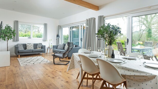 A spacious living area with a dining table set for four, a grey sofa, a wooden ceiling, and large windows offering a view of the outdoors. - Little Dunley Cottages - Apple Lodge (Bovey Tracey)