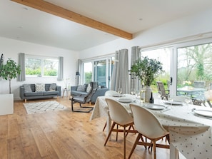 Een ruime woonkamer met een eettafel voor vier personen, een grijze bank, een houten plafond en grote ramen met uitzicht op het landschap.