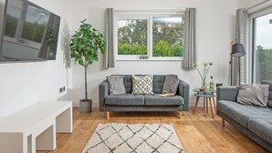 A modern living room with a grey sofa, a white bench, a potted plant, a window with curtains, and a floor lamp. - Little Dunley Cottages - Apple Lodge (Bovey Tracey)