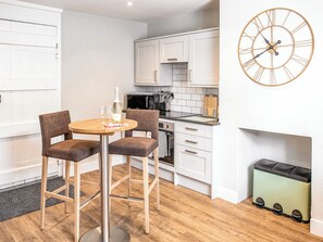 A modern kitchen with white cabinets, a wooden dining table with two chairs, a wall clock, and a trash bin.