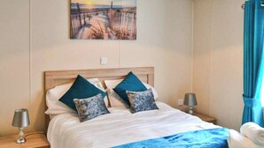 Quarto com cabeceira de madeira, duas mesas de cabeceira com luminárias, colcha azul e uma pintura de uma paisagem marítima acima da cama.