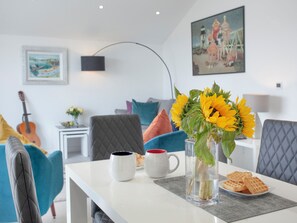 Ein modernes Wohnzimmer mit einem weißen Tisch, blauen Stühlen, einer Vase mit Sonnenblumen und einer Gitarre im Hintergrund.