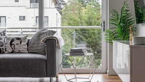 Een moderne woonkamer met een grijze bank, een klein bijzettafeltje en uitzicht op een balkon met groen.