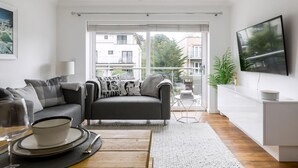 Een moderne woonkamer met een bank, een eettafel, een televisie en een balkon met uitzicht op gebouwen.