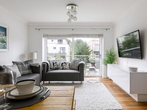 A modern living room with a sofa, a dining table set, a TV, and a balcony with a view of buildings. - 3 bedroom accommodation in Bournemouth (Bournemouth)