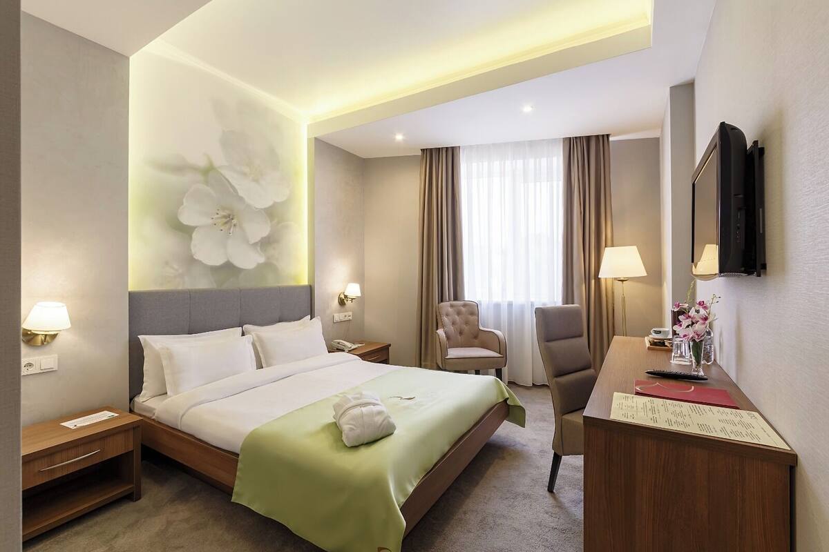 A hotel room with a large bed, a desk, a chair, a TV, and a wall-mounted lamp.