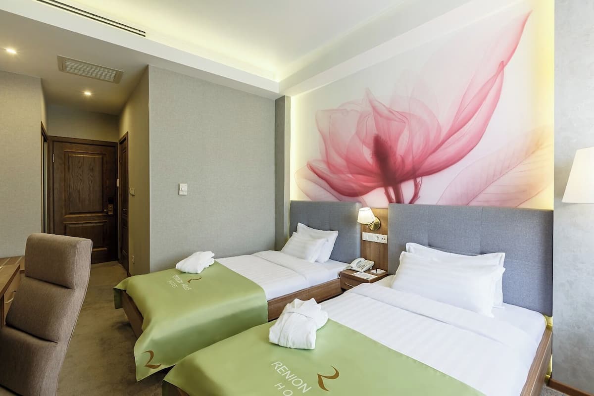 A hotel room with two beds, a chair, a large floral wall mural, and a bedside table with a lamp.