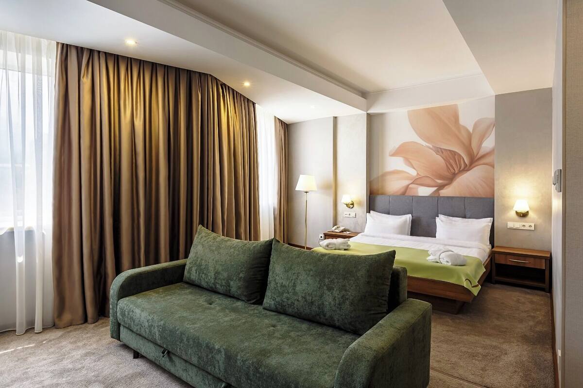 A hotel room with a green sofa, a bed with white linens, a large floral wall mural, and a floor lamp.