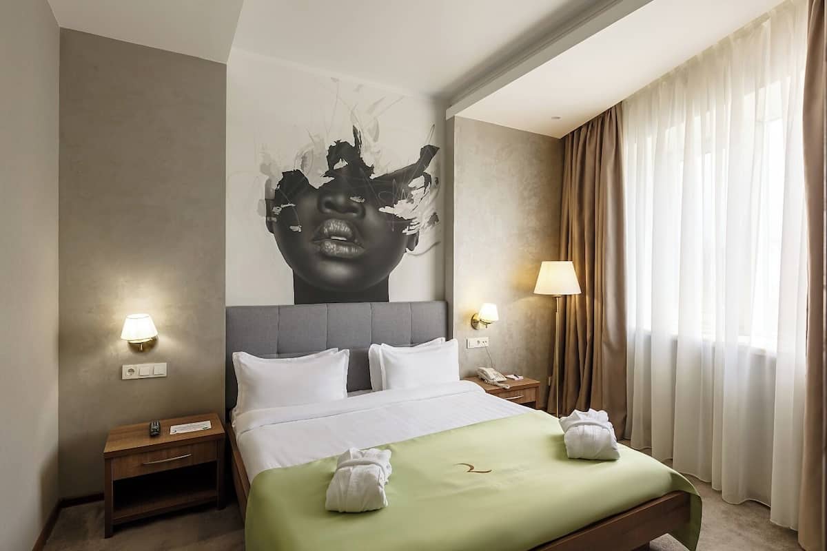 A hotel room with a large bed, two bedside tables, a nightstand, a wall-mounted lamp, and a large window with curtains.