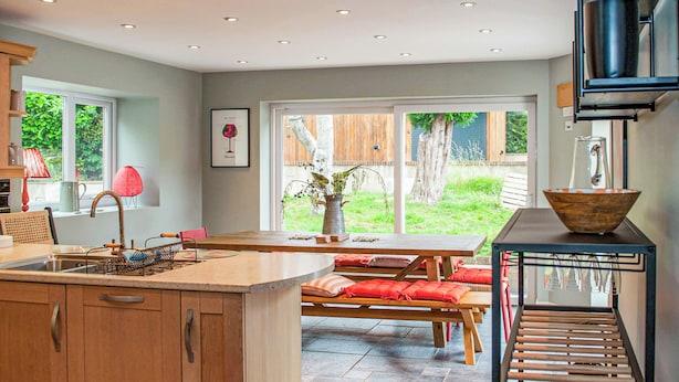 A modern kitchen with a central island, wooden cabinets, and a dining area with a bench and red cushions. - 4 bedroom accommodation in Uplyme (Uplyme)
