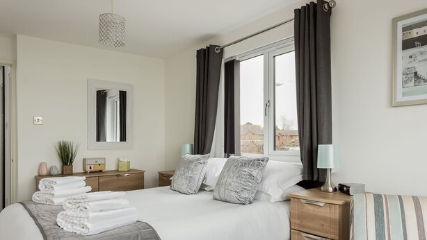 Een net opgemaakt bed met een grijze dekbedovertrek, twee kussens en een bedlampje. Er staat een houten nachtkastje met een lamp en een klein plantje. Een raam met gordijnen is zichtbaar, en aan de muur hangt een lijst met een foto.