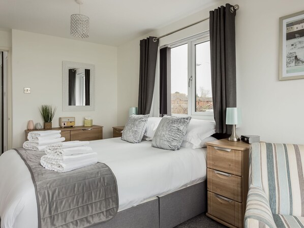 A neatly made bed with a grey bedspread, two pillows, and a bedside lamp. There is a wooden nightstand with a lamp and a small plant. A window with curtains is visible, and a framed picture hangs on the wall. - 2 bedroom accommodation in Poole (Poole)
